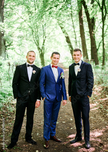 Groom in bright blue suit and gromsmen in black tuxedo stand on path in the forest