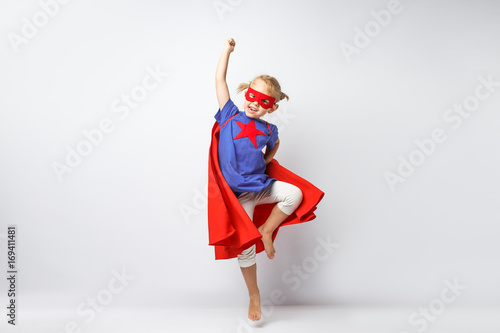 Very excited little girl dressed like superhero jumping alongside the white wall Fototapet