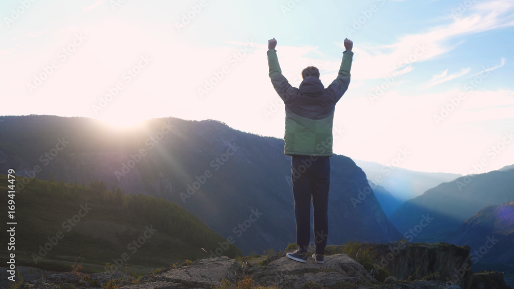 Young caucasian man raising his hands high on top of the big mountain at beautiful golden sunset Young caucasian man raising his hands high on top of the big mountain at beautiful golden sunset