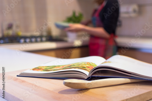 Cook book put on a kitchen table with a wooden spoon in the foreground. A young woman with apron is cooking in the Kitchen. © Sabrina