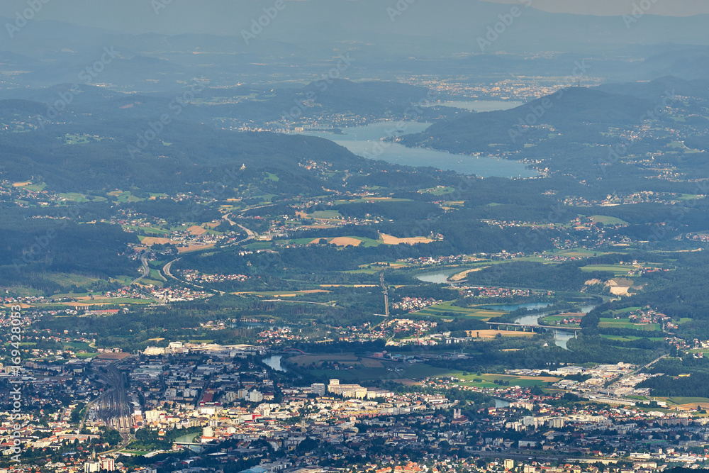 Aerial view of Villach City center with surrounding roads and railway tracks and with Wörther See in background covered in haze