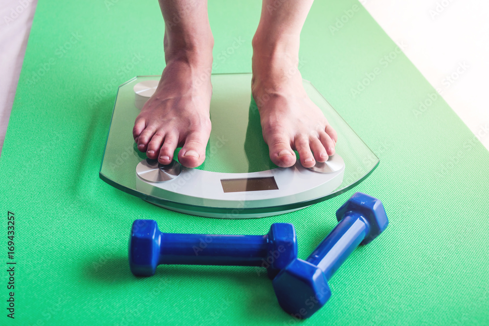 Fototapeta premium Female feet standing on electronic scales, dumbbells. Concept of sports training, slimming and weight loss