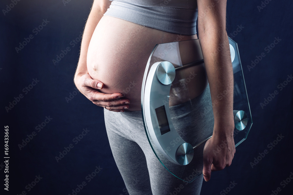 Pregnant woman holding scales and weigh keeping one hand on a belly. Concept of weight loss and control during pregnancy - obrazy, fototapety, plakaty 