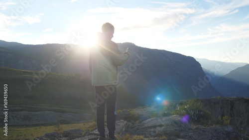 Tourist man making photos with smart phone on peak of rock in the mountains with lens flare effects.