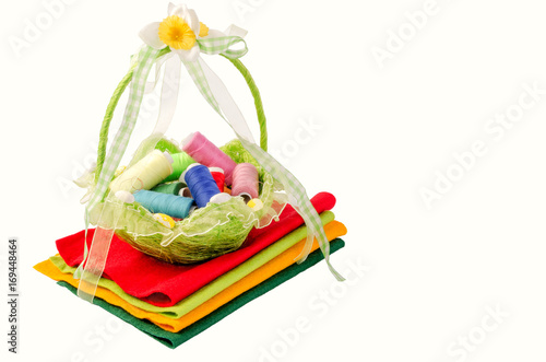 A beautiful wicker basket with multi-colored spools of thread stands on multi-colored fabric pieces. Isolated on white background.