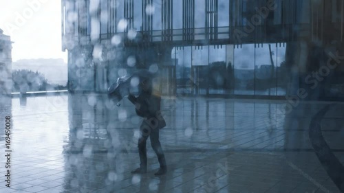 Young businessman in suit with umbrella under rain. Business centre building at the background. View through window with drops of water. Young man is running with an umbrella in the rain