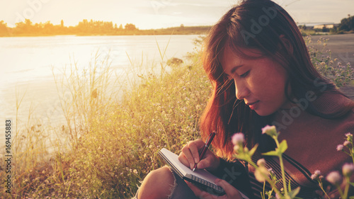 Young woman with pen writing on notebook at riverside in the evening.