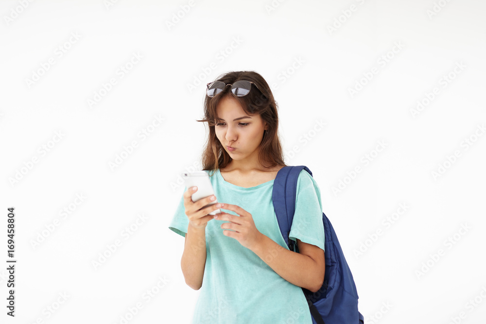 Horizontal picture of cute casually dressed mixed race female student carrying shoulder bag, reading text message on her generic smart phone with confused thoughtful expression on her cute face Horizontal picture of cute casually dressed mixed race female student carrying shoulder bag, reading text message on her generic smart phone with confused thoughtful expression on her cute face