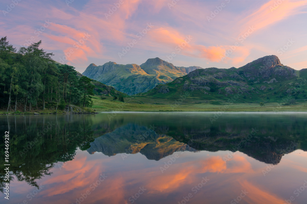Fototapeta premium Lustrzane odbicia w jeziorze o wschodzie słońca z porannym światłem uderzającym w góry w Lake District.