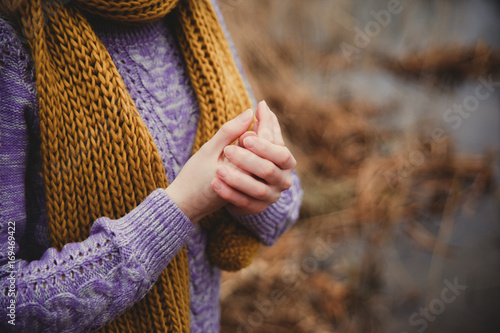 Fall sadness background. Unrecognisible girl in lilac knitted sweater and ocher scarf warming her hands close up.