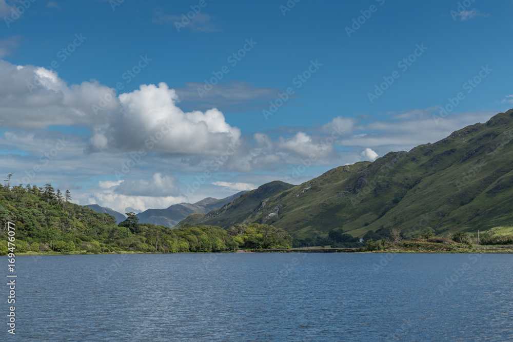 Connemara, Ireland - August 4, 2017: Looking East over Pollacapall Lough from the grounds of Kylemore Abbeys shows blue water, a dam, blue sky with white clouds and forested green hills.
