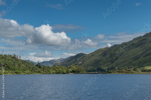 Connemara, Ireland - August 4, 2017: Looking East over Pollacapall Lough from the grounds of Kylemore Abbeys shows blue water, a dam, blue sky with white clouds and forested green hills. © Klodien