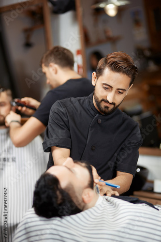 Handsome hairstylist in barber shop cutting bread with razor for brunet man.