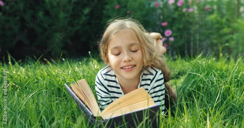 Portrait girl reading book in the grass and smiling at camera. Dolly shot Portrait girl reading book in the grass and smiling at camera. Dolly shot