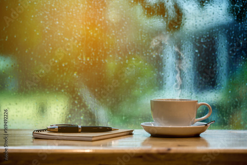 Cup of coffee, notebook and pen on wooden table on rainy day.