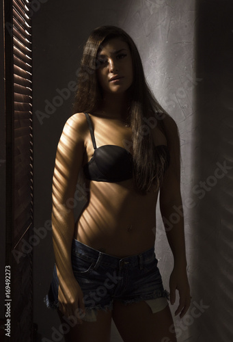 Beautiful woman in underwear stending near window and closed louver on a sunny day. Striped shadow on a body