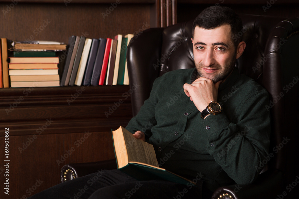Handsome the man with the book sits by bookshelves in a room with classic interior. Handsome the man with the book sits by bookshelves in a room with classic interior.