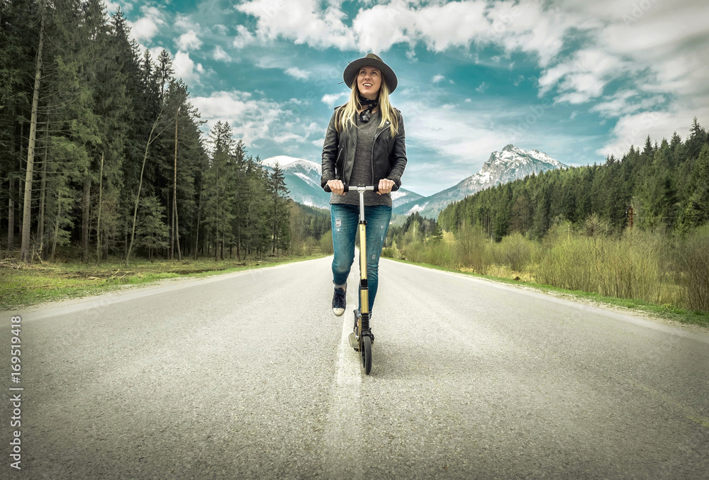 Obraz premium Woman on her kick scooter on the road around mountains, under s