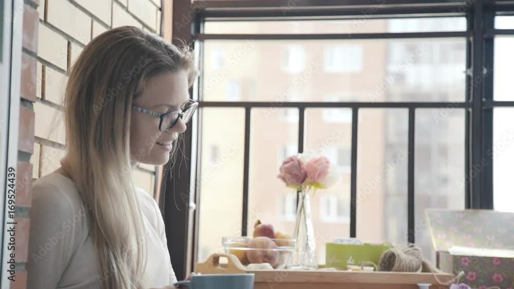 Modern young woman drinks coffee in the morning on a cozy balcony, tablet computer on her knees. Blonde woman with glasses holds cup of coffee and uses a tab (social networks, online shopping or work) Modern young woman drinks coffee in the morning on a cozy balcony, tablet computer on her knees. Blonde woman with glasses holds cup of coffee and uses a tab (social networks, online shopping or work)