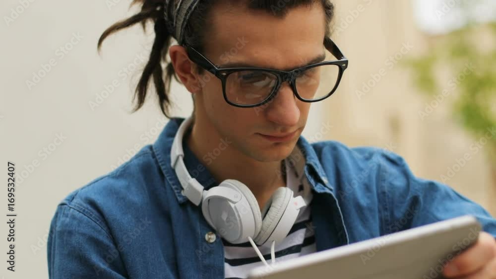 Portrait shot of young hipster man in the glasses typing in the tablet during resting in the city bench. Portrait shot of young hipster man in the glasses typing in the tablet during resting in the city bench.
