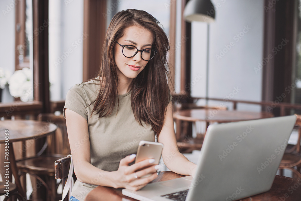 Beautiful woman using laptop and smartphone in a cafe. Young girl sitting in a coffee shop and working on computer Beautiful woman using laptop and smartphone in a cafe. Young girl sitting in a coffee shop and working on computer