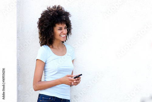 Smiling woman holding smart phone standing by white wall