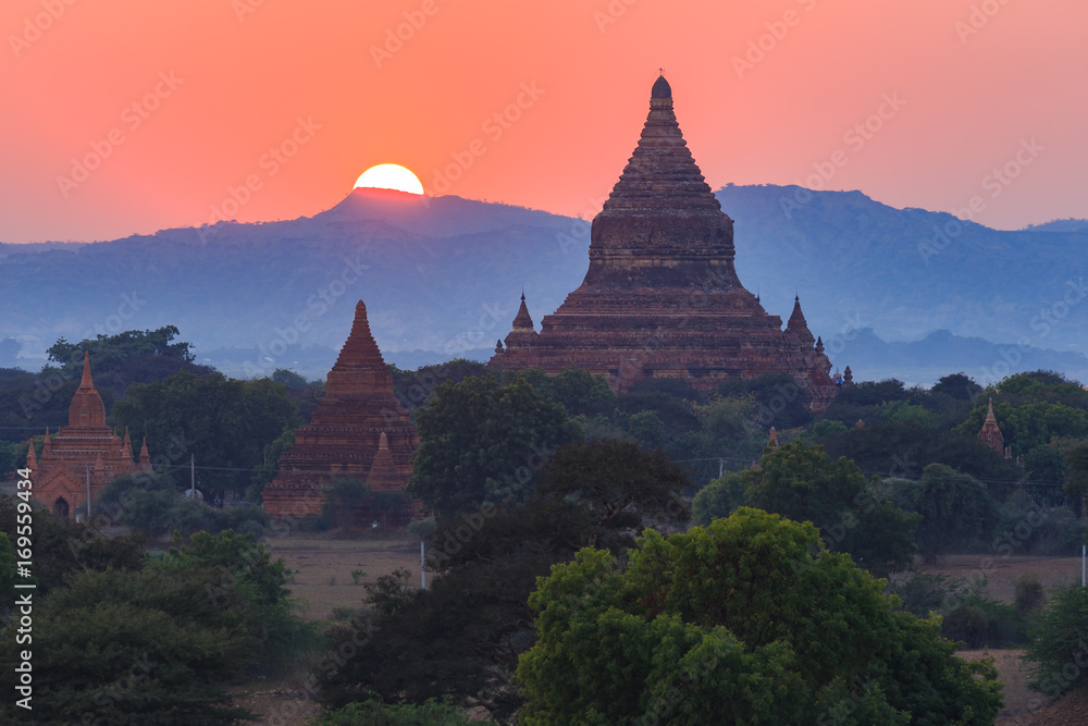 View from afar of the ancient pagodas (stupas) visible among rugged fields and trees of other pagodas and mountains on the horizon during sunset or sunrise, in Bagan, Myanmar (Burma)