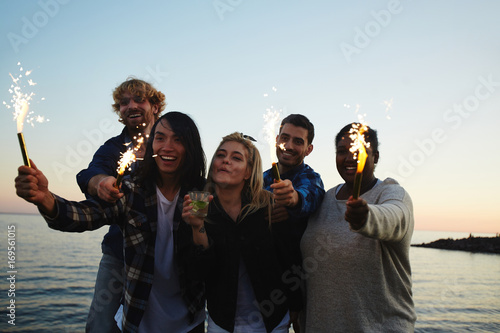 Joyful group of friends playing with sparklers while having fun on beach at sunset, pretty blonde woman drinking mojito cocktail, picturesque seascape on background