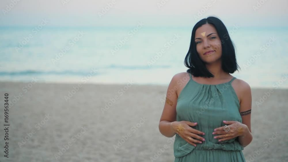 Outdoor fashion portrait of beautiful brunette lady at beach with flash tattoos on hands, back and forehead. Gypsy boho style. Woman in long green dress looking at camera. Slow motion. Outdoor fashion portrait of beautiful brunette lady at beach with flash tattoos on hands, back and forehead. Gypsy boho style. Woman in long green dress looking at camera. Slow motion.