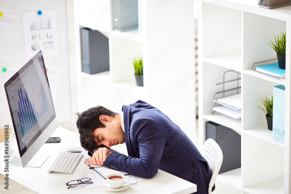 Overworked and tired young manager in stylish suit sleeping on desk in front of computer, interior of modern office on background Overworked and tired young manager in stylish suit sleeping on desk in front of computer, interior of modern office on background