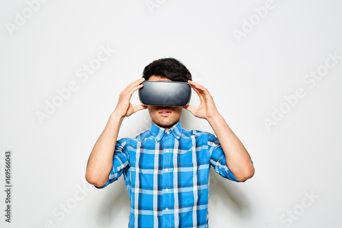 Waist-up portrait of young man wearing blue checked shirt using VR headset while standing against white background