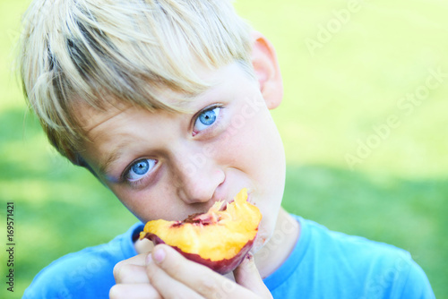 Portrait of child boy eating peach. Happy child in sun summer day. Kid with fruit in nature background.
