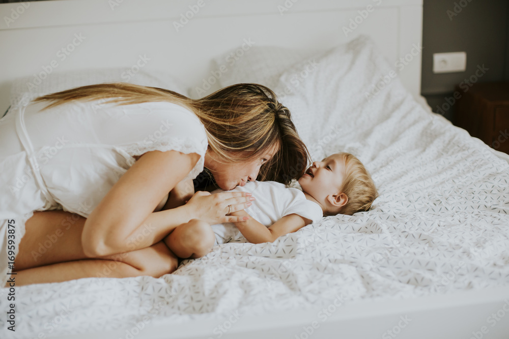 Young happy mother playing in a white bed enjoying a sunny morning in a modern home bedroom Young happy mother playing in a white bed enjoying a sunny morning in a modern home bedroom