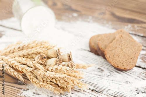 One bundle of wheat and poppy and flour poured out of glass and slices of bread on old rustic wooden planks