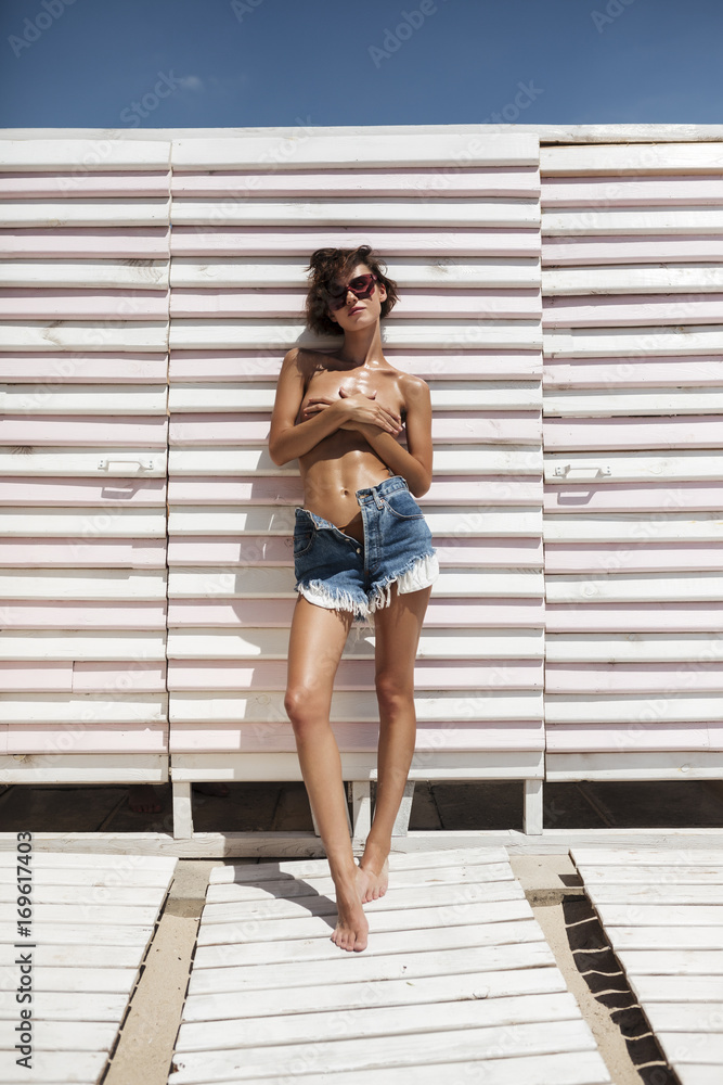 Obraz premium Portrait of beautiful girl in denim shorts and sunglasses spending time on beach. Young pretty girl standing and covering her chest with hands on beach