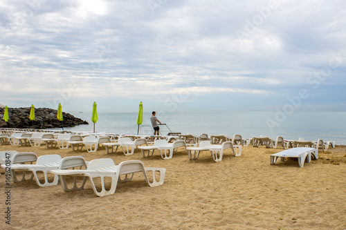 Man clean empty beach with rows of sun beds under straw umbrellas © sosiukin