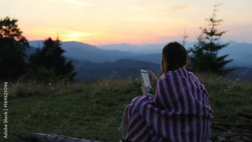 Young woman sitting using holding mobile smart phone wrapped blanket evening sunset setting sun horizon colorful sky high peaks mountains digital texting social network online tapping back view Young woman sitting using holding mobile smart phone wrapped blanket evening sunset setting sun horizon colorful sky high peaks mountains digital texting social network online tapping back view