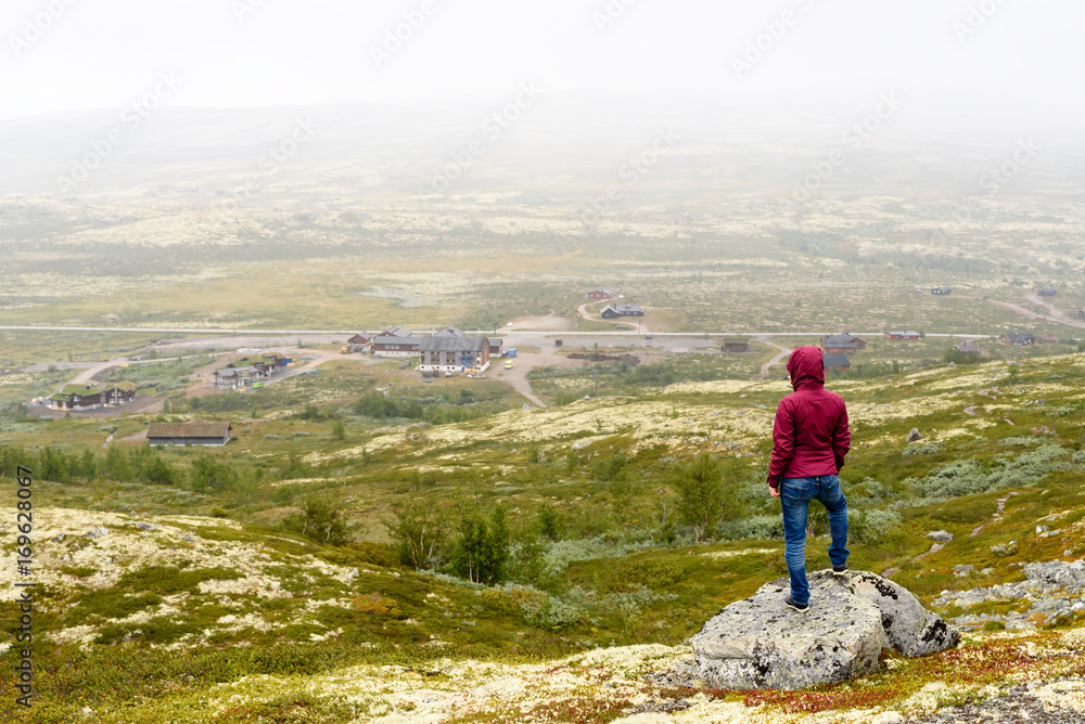 Young adult person looking out over foggy mountain view with small village and road. Location Vasstulan, Norway.