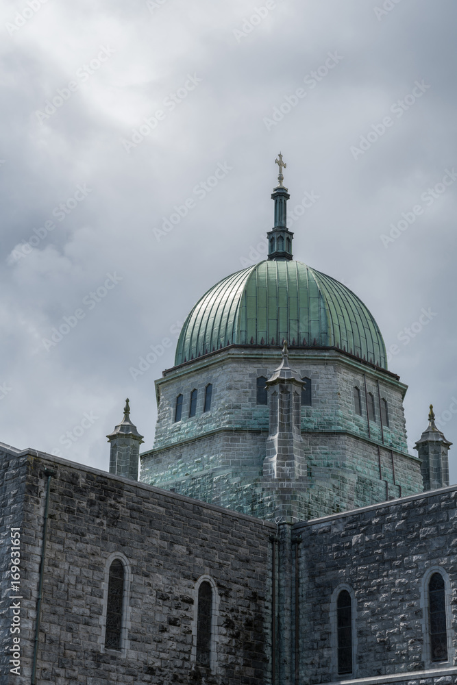 Galway, Ireland - August 5, 2017: Outside view of green dome with cross of the Cathedral under dark skies. Gray stone walls of the church.