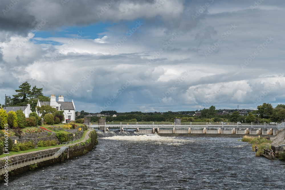 Galway, Ireland - August 5, 2017: Speed and volume of the fast flowing Corrib River is controlled by a dam system with floodgates upstream of the city. Cloudscape, trees, white water and 1899 office.