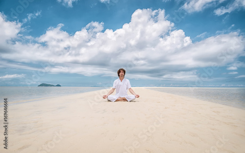 Young man meditating in lotus position on the beautiful long beach with clouds background.