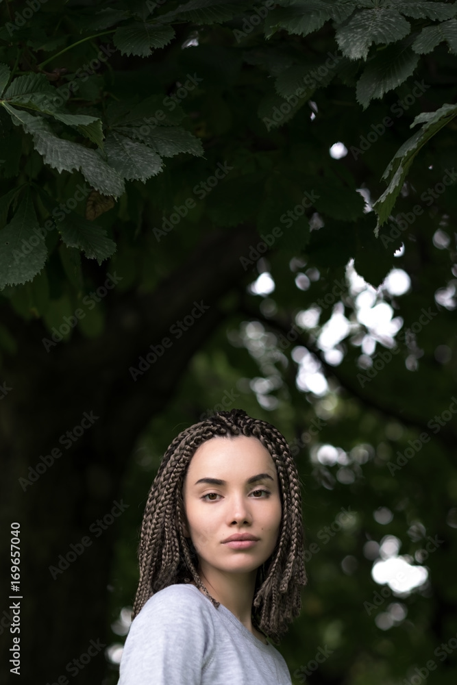 A beautiful girl with afro pigtails seriously looks at the camera on a background of green foliage of trees. A beautiful girl with afro pigtails seriously looks at the camera on a background of green foliage of trees.