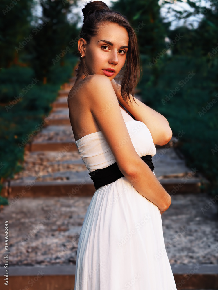 Young brunette model at the white dress staying at the stairs with green trees and looking into camera