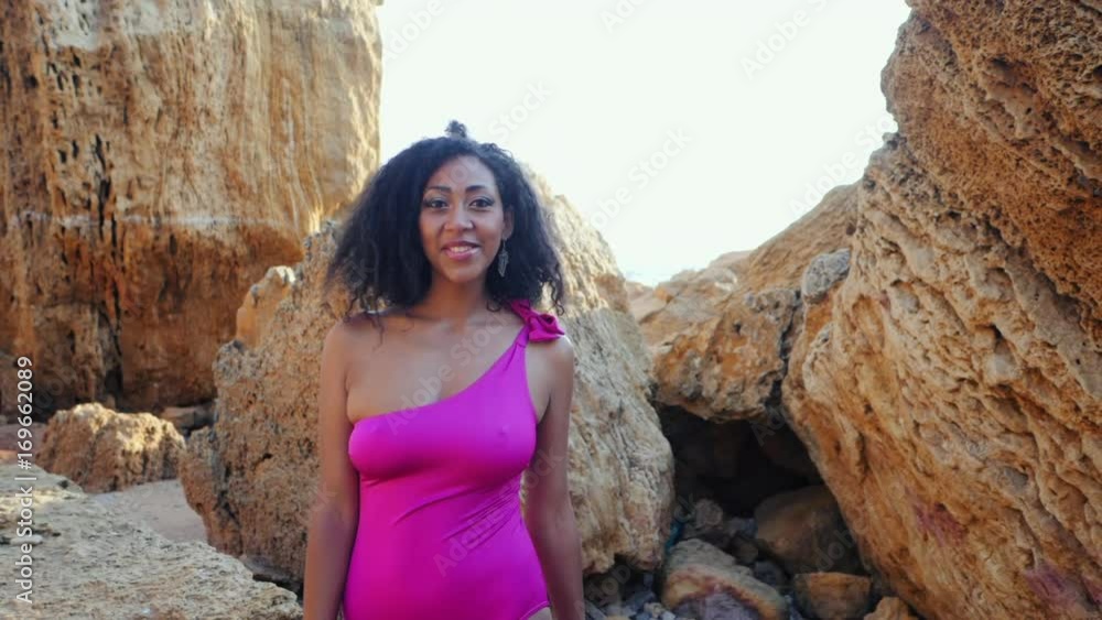 Portrait of black people, pretty happy young african american woman looking at camera and smiling.Rocks beach.Sexy mixed race girl in pink swimsuit and jewelry enjoying nature.Slow motion. Portrait of black people, pretty happy young african american woman looking at camera and smiling.Rocks beach.Sexy mixed race girl in pink swimsuit and jewelry enjoying nature.Slow motion.
