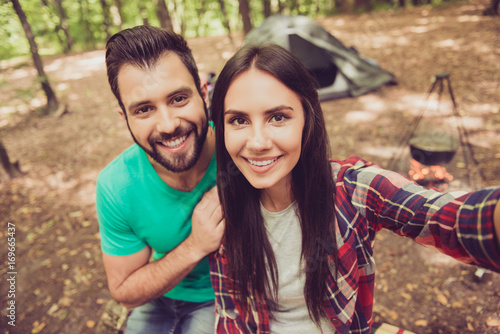 Close up shot of two young cheerful lovers in the summer nice wood, embracing, posing for a selfie shot, that beautiful brunette lady is taking