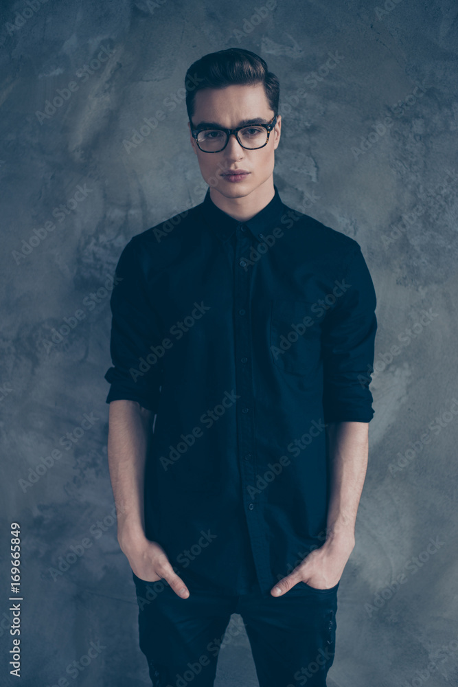 Vertical photo of thoughtful young stylish guy in trendy clothes and spectacles holding his hands in pockets