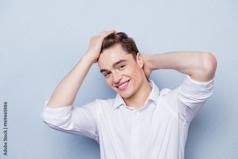 Portrait of happy with beaming smile young guy dressed in white shirt with rolled sleeves posing against gray background and touching his hair Portrait of happy with beaming smile young guy dressed in white shirt with rolled sleeves posing against gray background and touching his hair