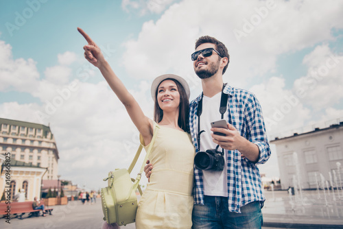 Check this out! Happy young couple of tourists are on vacation, in casual outfits, cap, eyewear and with camera, outdoors, nice spring day © deagreez