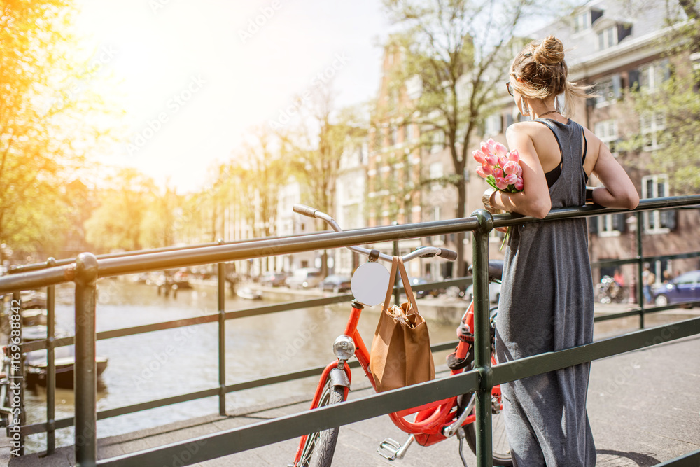 Fototapeta premium Młoda i piękna kobieta stojąca z rowerem i kwiatami na moście nad kanałem wodnym w mieście Amsterdam