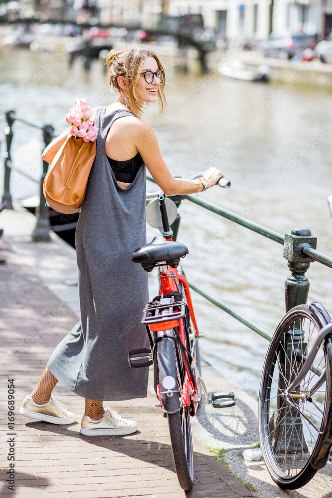 Fototapeta premium Młoda i piękna kobieta z torbą i kwiatami, spacery z rowerem na kanale wodnym w mieście Amsterdam
